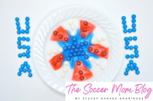 Red, white, and blue skittles arranged in a firework pattern on a white plate, with the colors dissolving in water.
