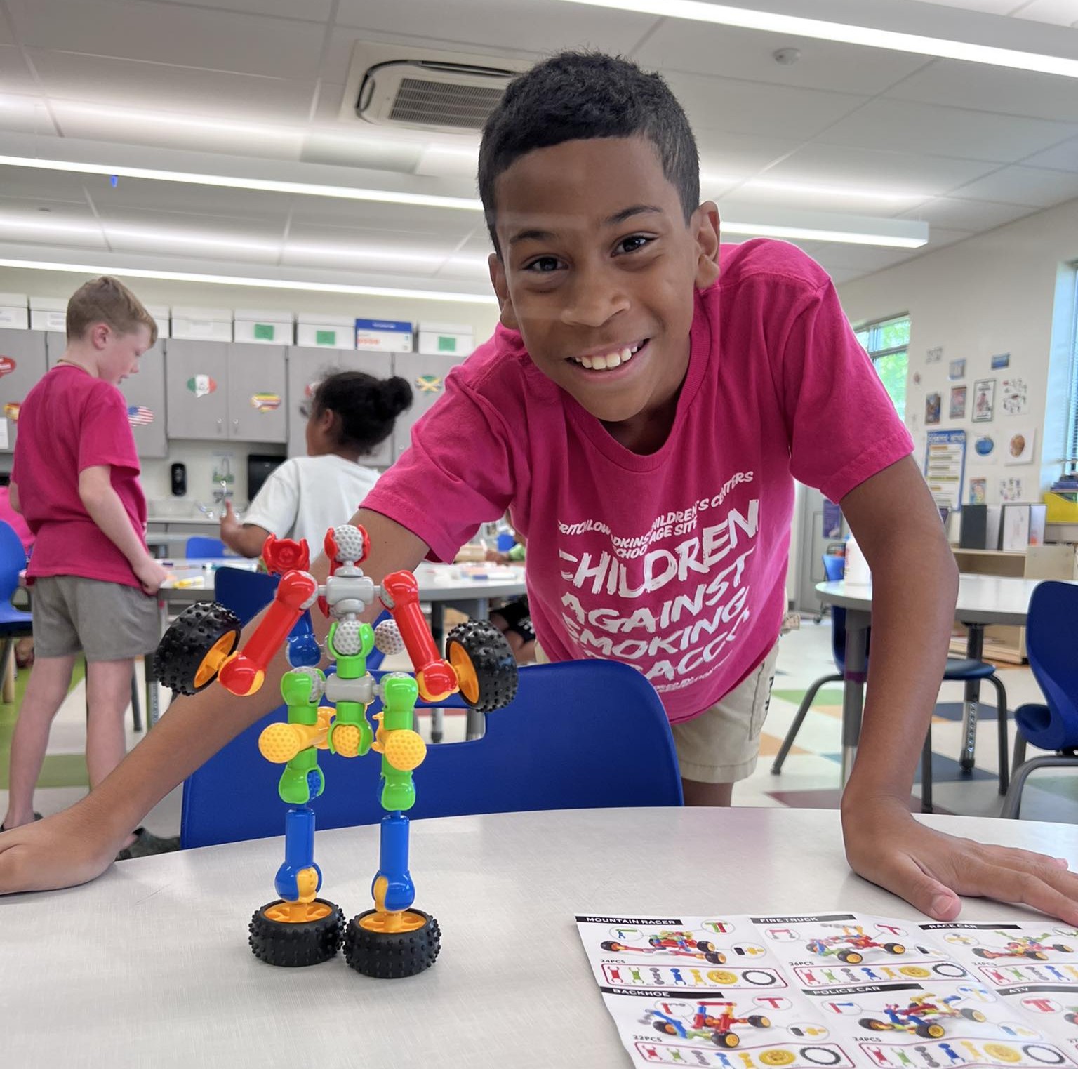 529766208_25054440424146701_2113038480859863088_n Boy in a classroom smiling with a robot he built sitting on the desk in front of him.